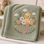 May include: A sage green knitted blanket featuring an embroidered design of a yellow duckling surrounded by white and pink flowers, green leaves, and white clouds. The name Hazel is embroidered in pink script lettering below the duckling.