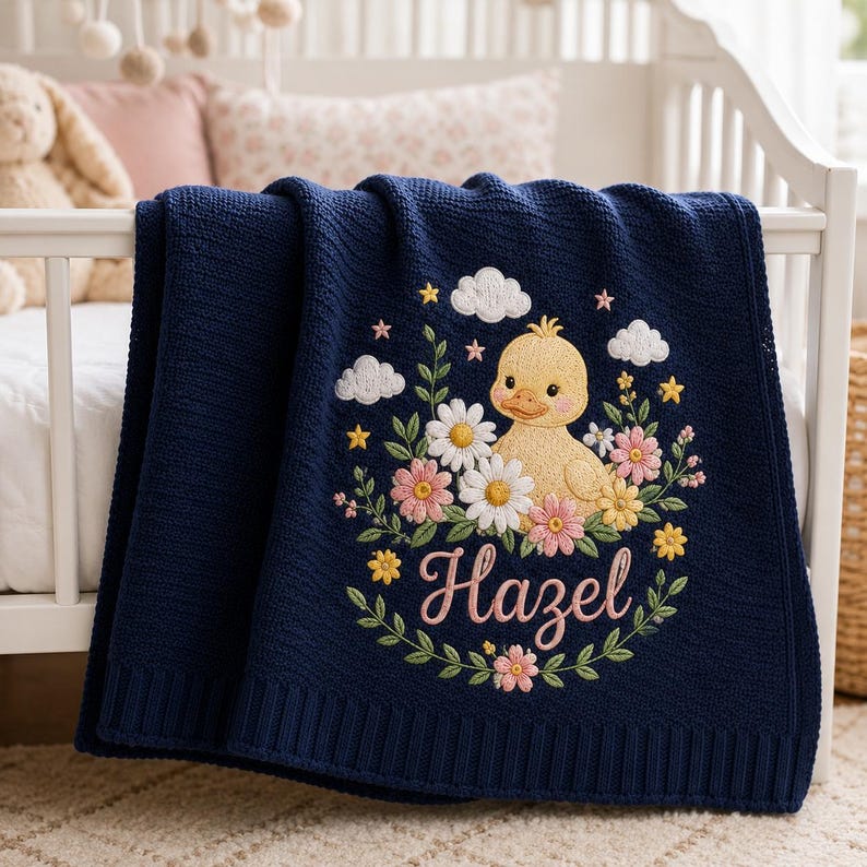 May include: A navy blue knit baby blanket featuring an embroidered design of a yellow duckling surrounded by white and pink flowers, green leaves, small yellow stars, and white clouds. The name Hazel is embroidered in pink script below the