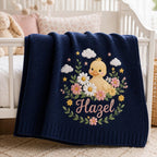 May include: A navy blue knit baby blanket featuring an embroidered design of a yellow duckling surrounded by white and pink flowers, green leaves, small yellow stars, and white clouds. The name Hazel is embroidered in pink script below the