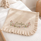 May include: A cream-colored knitted blanket featuring a ruffled edge and an embroidered floral wreath design surrounding the name Evelyn in green script. The blanket is made of a soft knit fabric with delicate flower details in pink, white, and blue