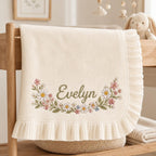 May include: A cream-colored knit baby blanket featuring a ruffled edge and an embroidered floral garland surrounding the name Evelyn in green script. The design includes small pink, white, and blue flowers with green leaves on a soft fabric.