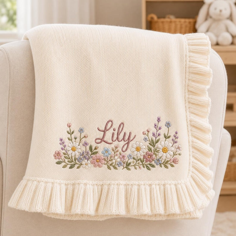 May include: A cream-colored knit baby blanket featuring a ruffled edge and an embroidered floral arrangement in shades of pink, blue, and white. The name Lily is embroidered in a cursive script in the center of the floral design.