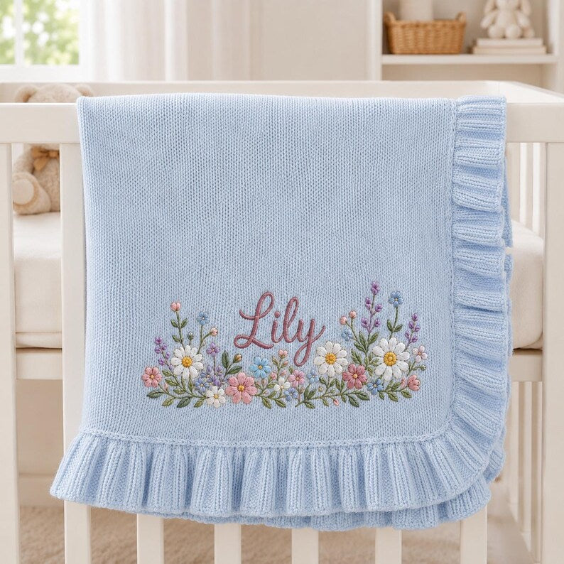 May include: A light blue knit baby blanket featuring a ruffled edge and an embroidered floral arrangement at the bottom. The name Lily is stitched in a dark pink script font in the center of the colorful flower design, which includes daisies.