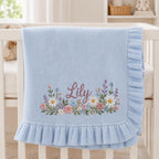 May include: A light blue knit baby blanket featuring a ruffled edge and an embroidered floral arrangement at the bottom. The name Lily is stitched in a dark pink script font in the center of the colorful flower design, which includes daisies.
