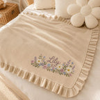 May include: A beige knit blanket featuring a ruffled border and an embroidered floral arrangement with the name Lily in cursive script. The design includes small white, purple, and blue flowers with green leaves centered on the fabric surface.