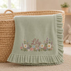 May include: A sage green knit baby blanket featuring a ruffled edge and an embroidered floral arrangement at the center. The name Lily is stitched in a cursive brown font above a colorful cluster of daisies, lavender, and small pink flowers.