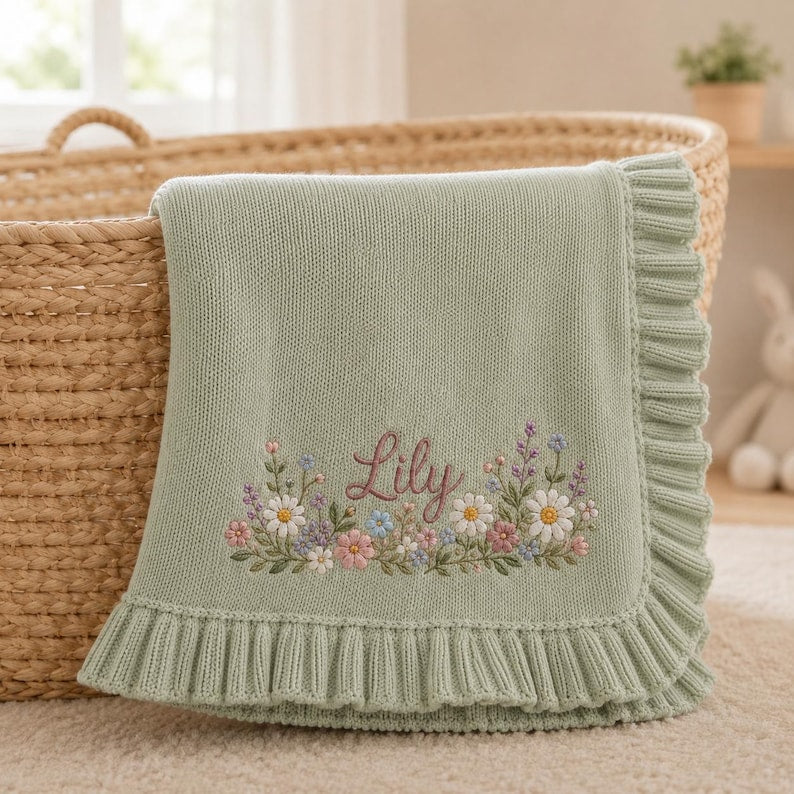 May include: A sage green knit baby blanket featuring a ruffled edge and an embroidered floral arrangement at the center. The name Lily is stitched in a cursive brown font above a colorful cluster of daisies, lavender, and small pink flowers.