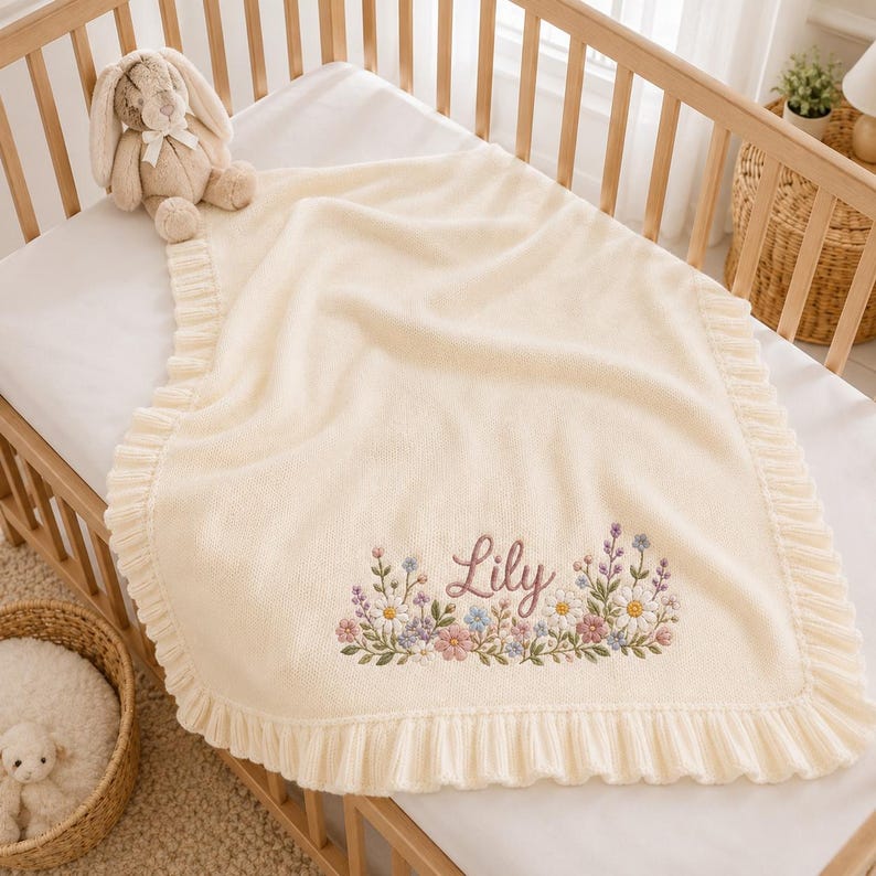 May include: A cream-colored knit baby blanket featuring a ruffled border and an embroidered floral arrangement at the bottom. The name Lily is stitched in cursive script above the flowers. A plush tan rabbit toy sits on the corner of the blanket.