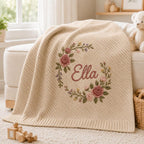May include: A beige knit blanket featuring a diagonal ribbed texture and a central embroidered floral wreath. The wreath is composed of pink roses, green leaves, and small purple and yellow accents. The name Ella is embroidered in cursive script.