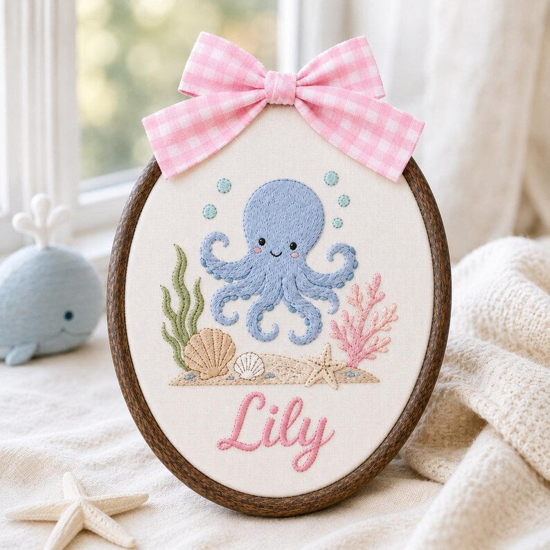 May include: An oval embroidery hoop featuring a light blue octopus, green seaweed, pink coral, and seashells stitched on white fabric. The name Lily is embroidered in pink script below the sea life. A pink and white gingham bow sits on top