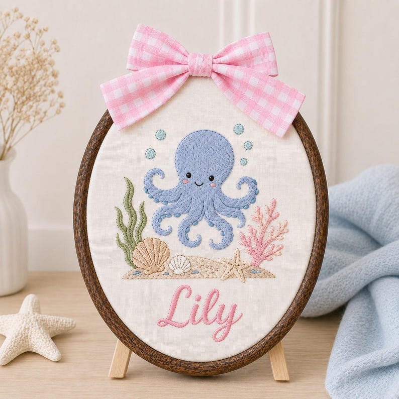 May include: An oval embroidery hoop containing a white fabric panel with a blue octopus, seashells, and coral stitched on it. The name Lily is embroidered in pink script below the sea life. A pink and white gingham bow is attached to the t