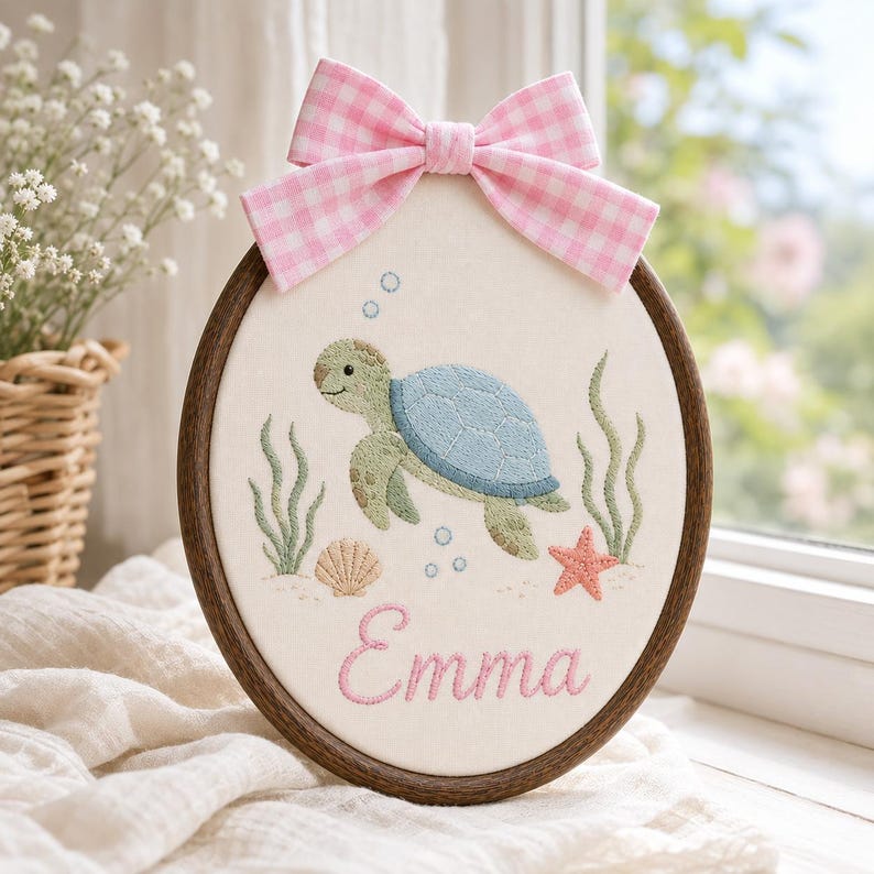 May include: An oval embroidery hoop containing a fabric panel with a stitched sea turtle, seaweed, a seashell, and a starfish. The name Emma is embroidered in pink script at the bottom. A pink and white gingham bow is attached to the top o