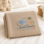 May include: A beige knit blanket featuring an embroidered design of a blue whale, a green sea turtle, coral, seashells, and a yellow starfish. The text "Sweet Dreams" is embroidered in blue script below the whale and turtle imagery.