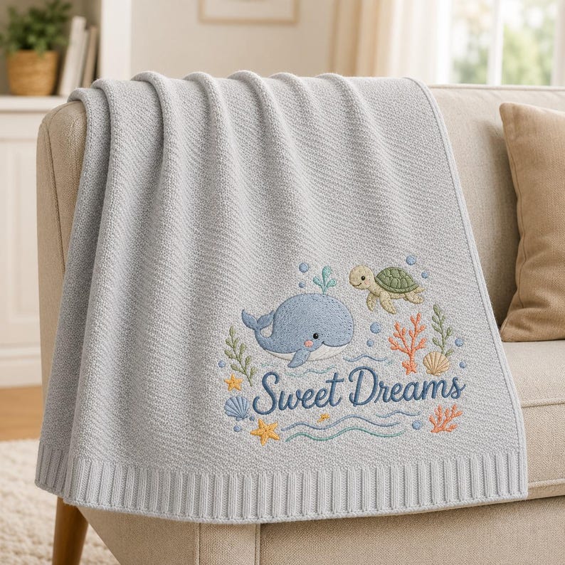 May include: A light gray knit blanket featuring an embroidered design of a blue whale, a green sea turtle, coral, seashells, and a yellow starfish. The text "Sweet Dreams" is embroidered in dark blue script below the sea creatures.