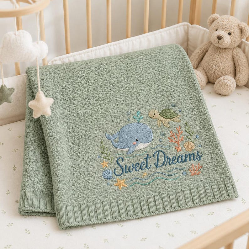 May include: A sage green knitted baby blanket featuring an embroidered design of a blue whale, a green sea turtle, coral, and seashells. The text "Sweet Dreams" is embroidered in dark blue script below the sea creatures on the textured fabric.