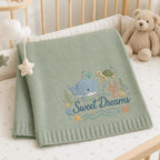 May include: A sage green knitted baby blanket featuring an embroidered design of a blue whale, a green sea turtle, coral, and seashells. The text "Sweet Dreams" is embroidered in dark blue script below the sea creatures on the textured fabric.