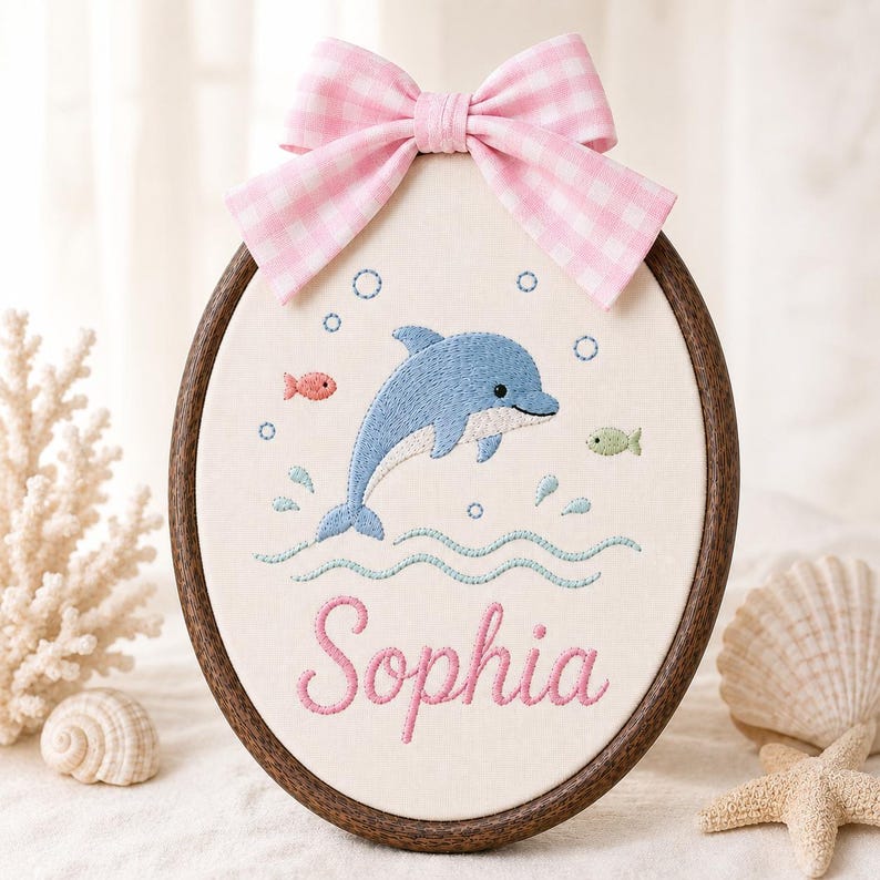 May include: An oval embroidery hoop featuring a blue dolphin, small fish, and bubbles stitched on off-white fabric. The name Sophia is embroidered in pink script below the dolphin. A pink and white gingham bow is attached to the top of the