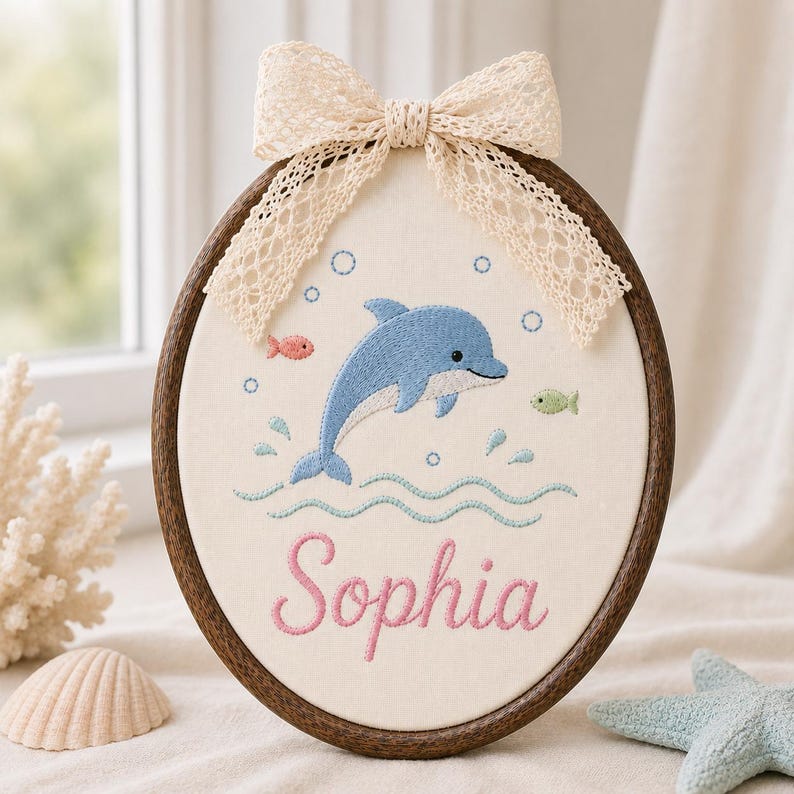 May include: An oval embroidery hoop frame containing cream fabric with a blue dolphin, small fish, and bubbles stitched in thread. The name Sophia is embroidered in pink script below the dolphin. A cream lace bow is attached to the top of 