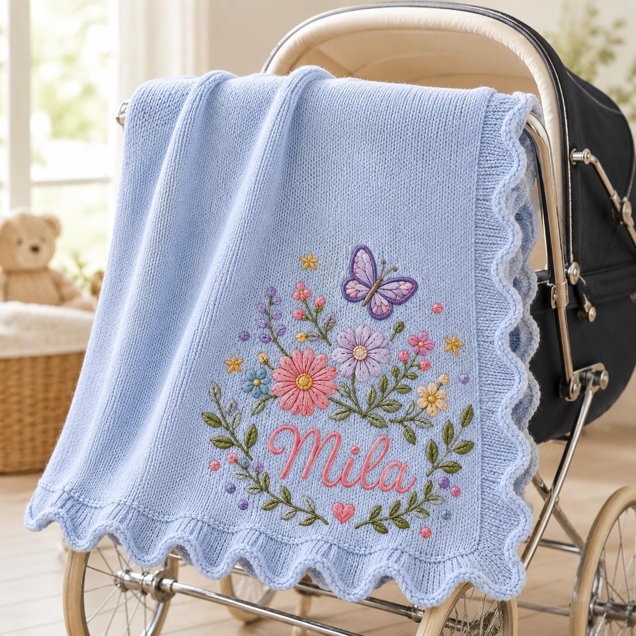 May include: A light blue knitted baby blanket with a scalloped edge, featuring an embroidered design of a purple butterfly, various colorful flowers, green leaves, and the name Mila in pink script lettering, draped over a dark-colored baby carriage.
