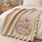 May include: A beige knitted baby blanket featuring a scalloped edge and an embroidered design of a purple butterfly, various colorful flowers, and green leaves. The name Mila is embroidered in pink cursive script in the center of the floral arrangem