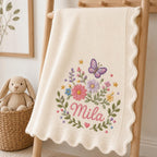 May include: A cream-colored knit baby blanket featuring a scalloped edge and an embroidered design of a purple butterfly, colorful flowers, green leaves, and small stars. The name Mila is embroidered in pink script in the center of the floral arrang