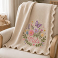 May include: A beige knit blanket featuring a scalloped edge and an embroidered design of a purple butterfly, various colorful flowers, and green leaves. The name Mila is embroidered in pink cursive script at the center of the floral arrangement.